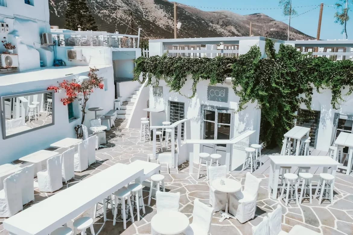 An elegant outdoor restaurant is filled with white tables and chairs, surrounded by traditional white buildings and lush greenery.