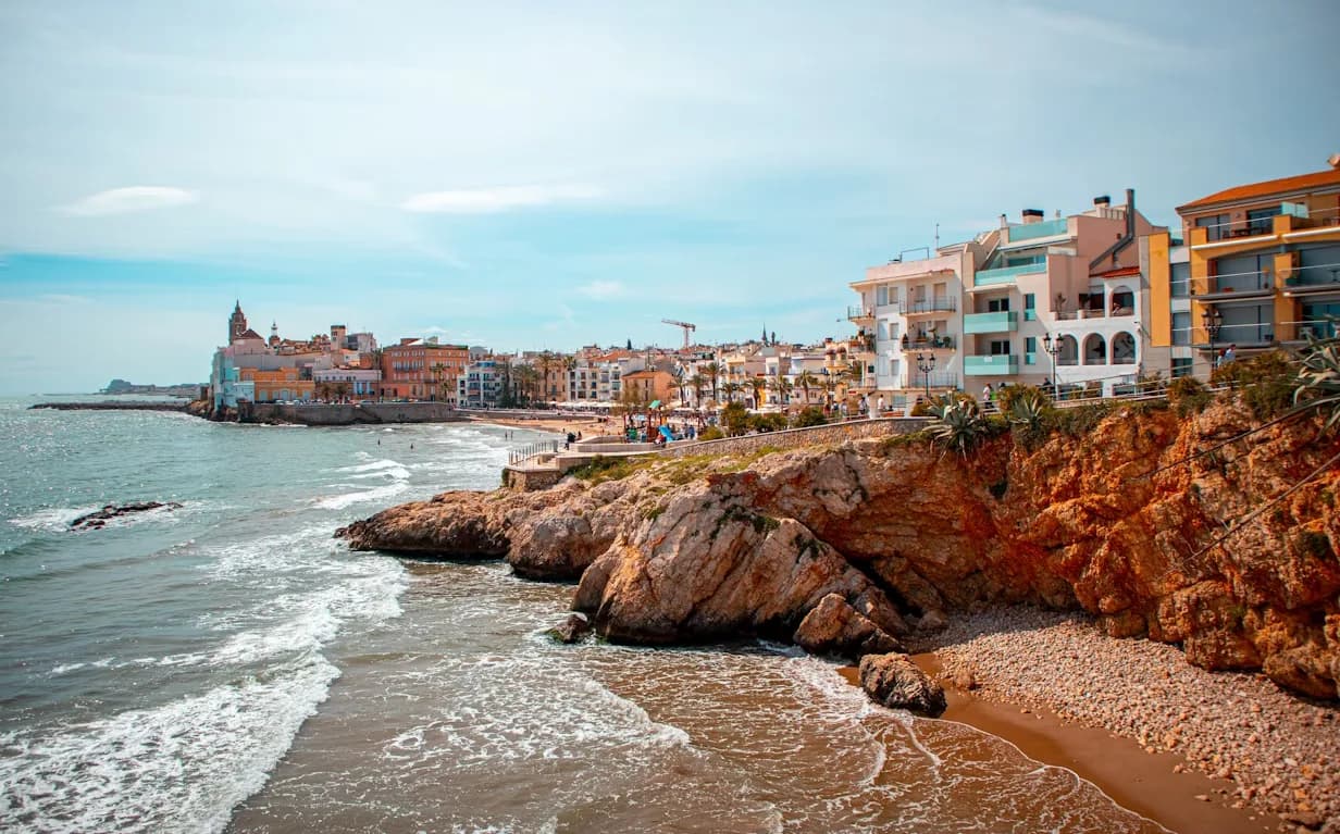 The historic town of Sitges is built on a rocky cliff overlooking the sea, with a series of buildings and a bell tower.