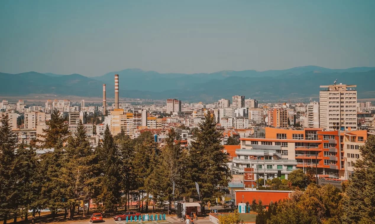 A panoramic view of the Sofia city skyline shows a mix of residential and modern buildings, with a large, forested mountain in the background.