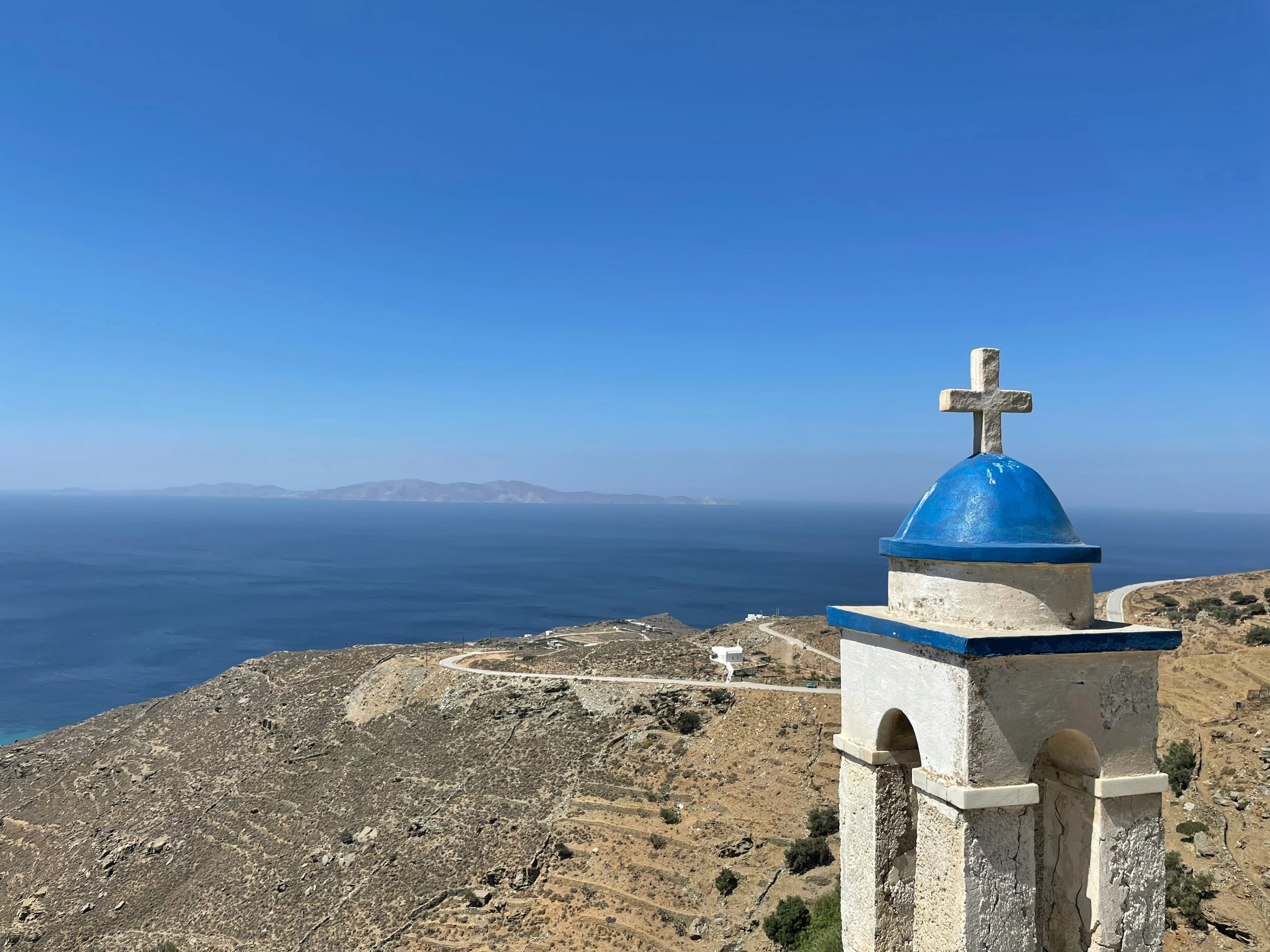 A beautiful, traditional Greek Orthodox church with a white facade and a bell tower stands on a hillside overlooking the Aegean Sea.