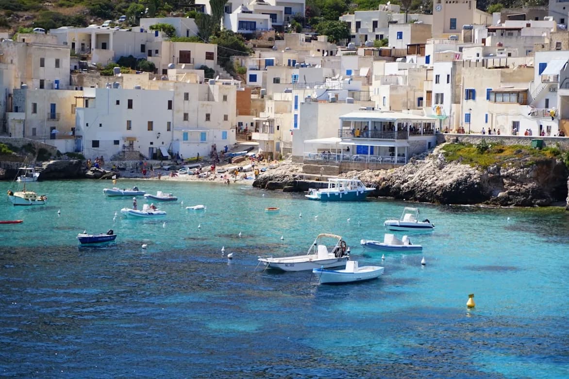 A wide, sandy beach is filled with people and colorful umbrellas, with the sea and the historic city of Trapani in the background.