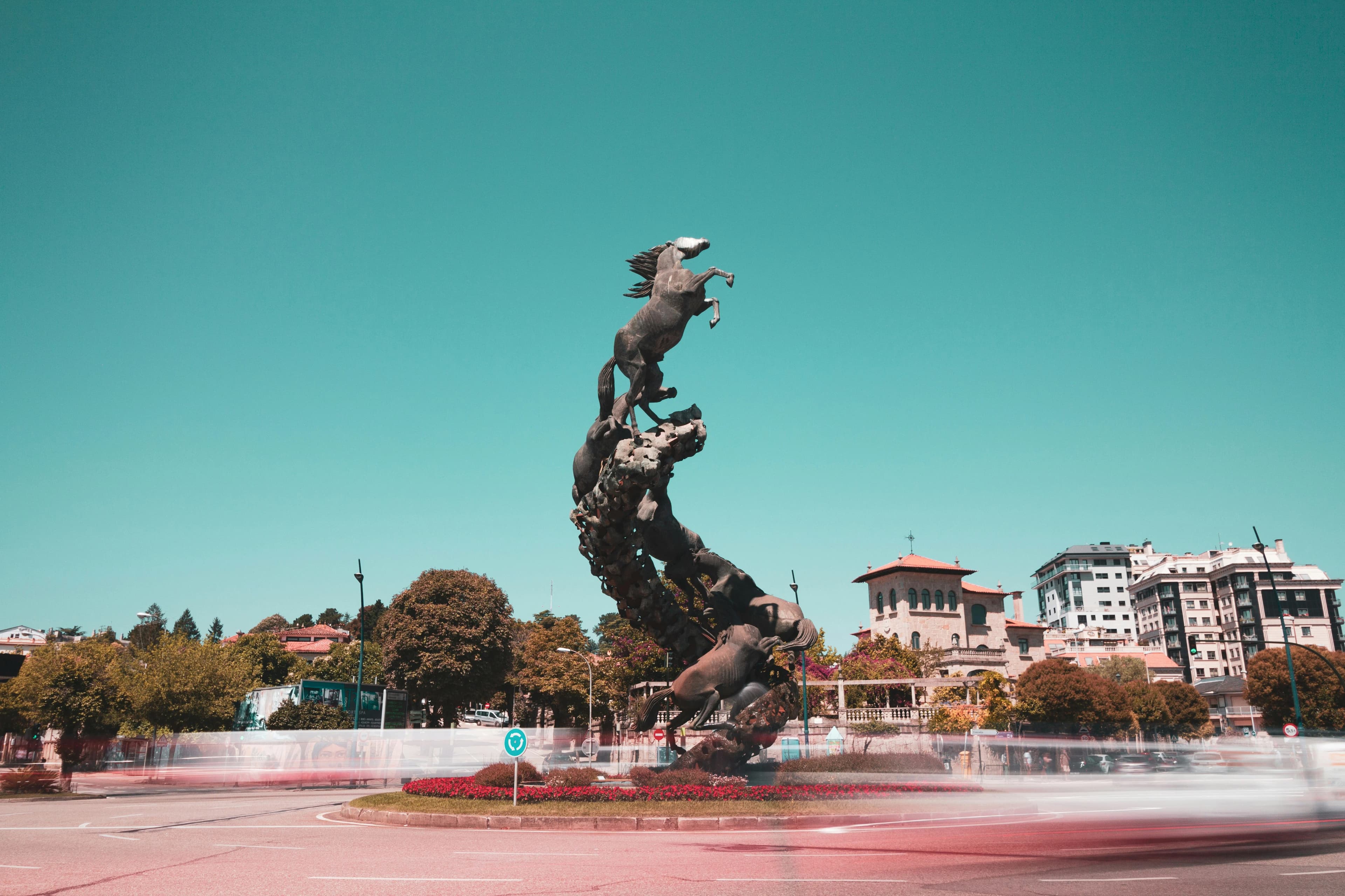 A large, ornate statue of several horses rearing up on a circular pedestal is a striking feature of a city roundabout.