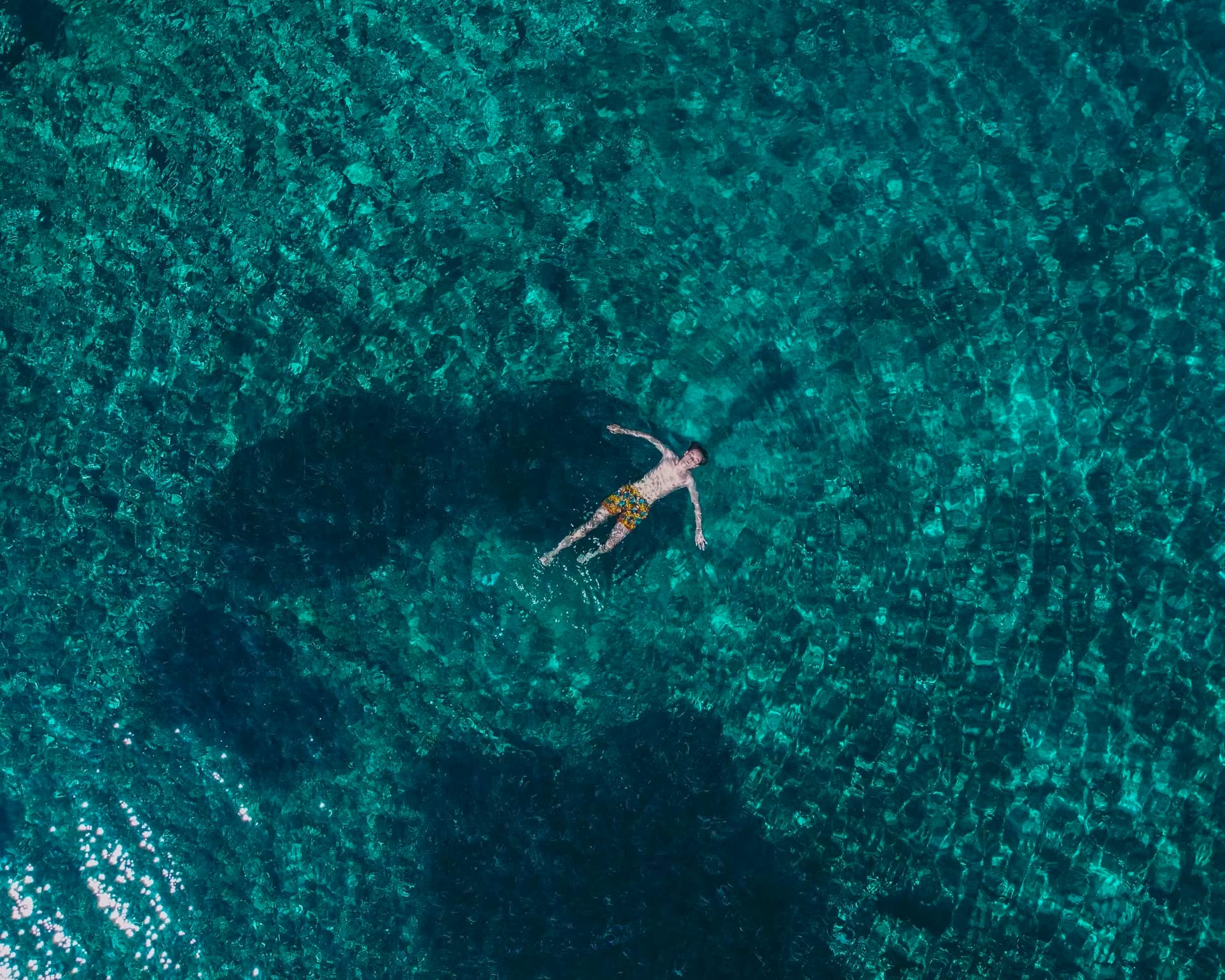 A person floats on their back in the remarkably clear, turquoise water, with sunlight creating a ripple effect on the surface.