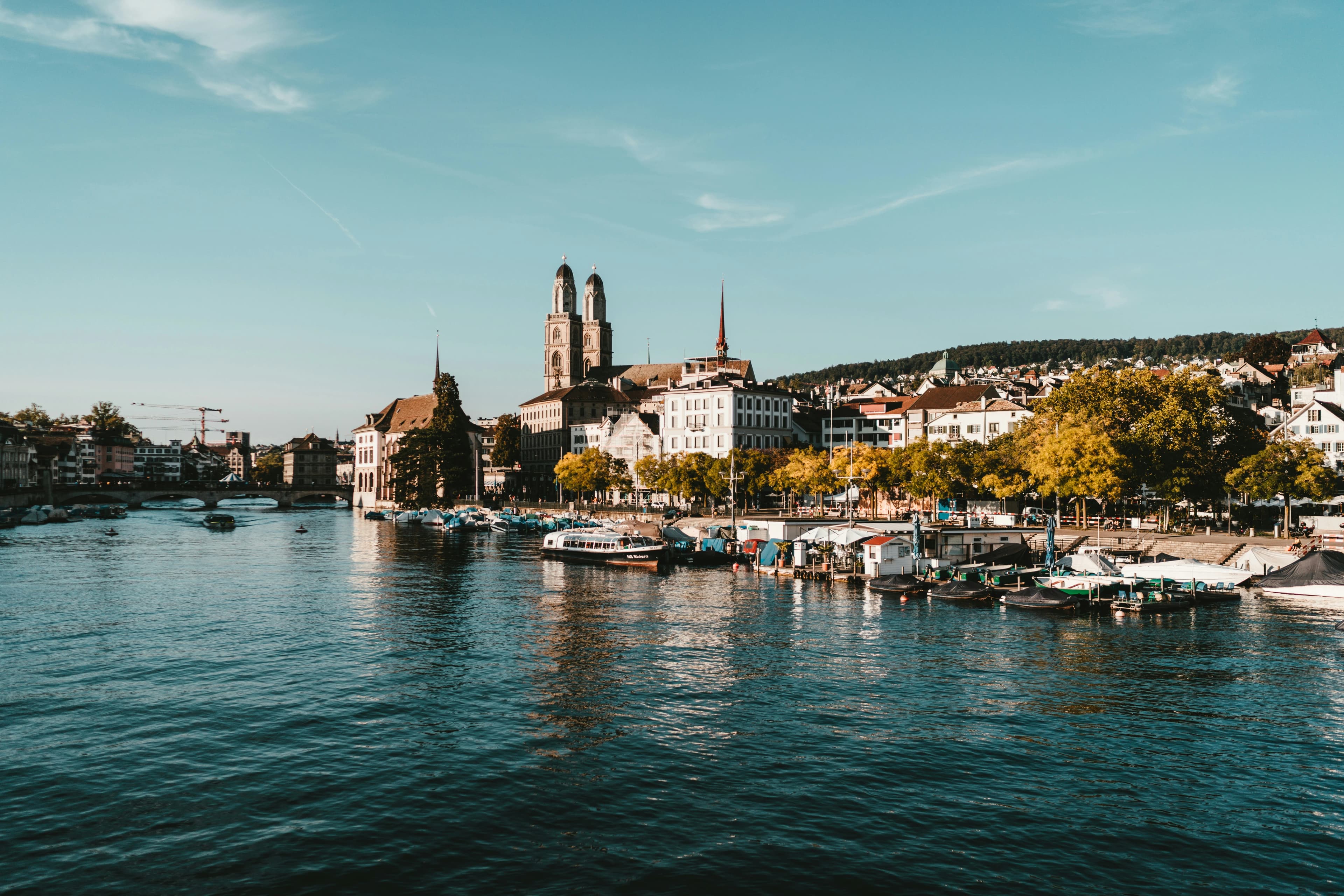 A daytime view of the Limmat River in Zurich, with city buildings and churches lining the banks.