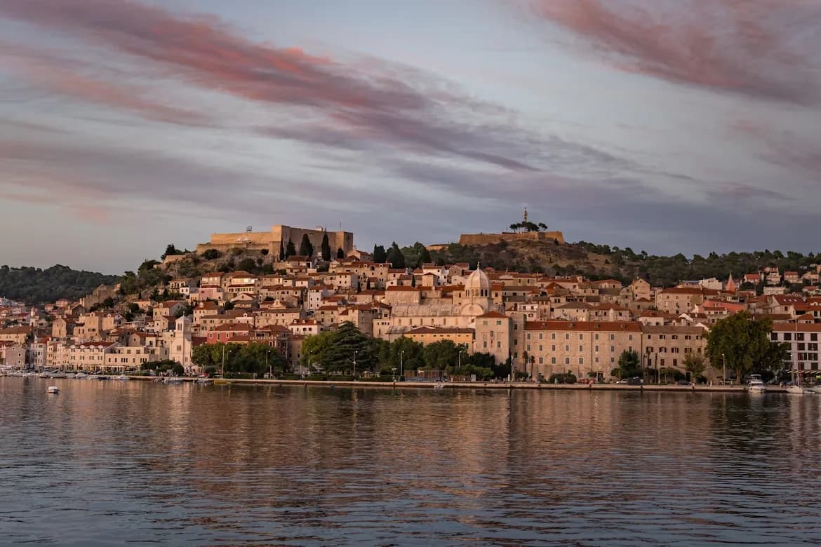 The historic city of Šibenik is beautifully illuminated by the setting sun, with the city's lights and a fortress on the hill reflecting on the sea.