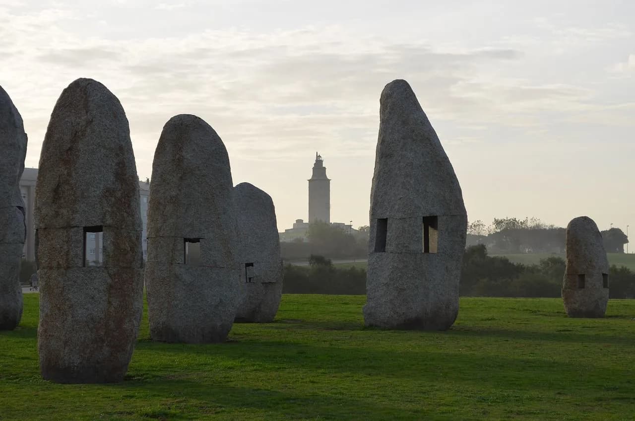 Ancient stone sculptures stand in a grassy field, with the historic Tower of Hercules lighthouse visible in the distance.