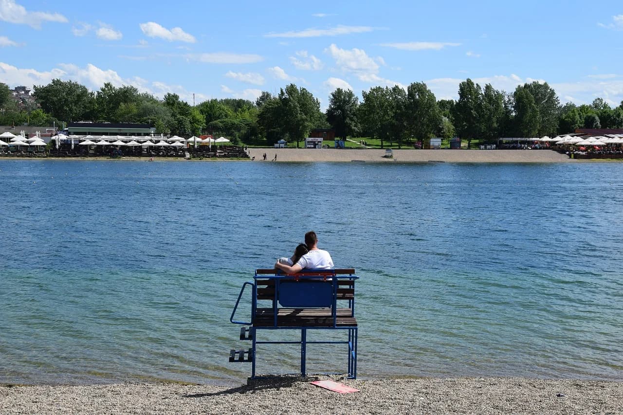 A couple sits on a wooden lifeguard stand, overlooking a tranquil lake and a sandy beach with cafes and umbrellas.