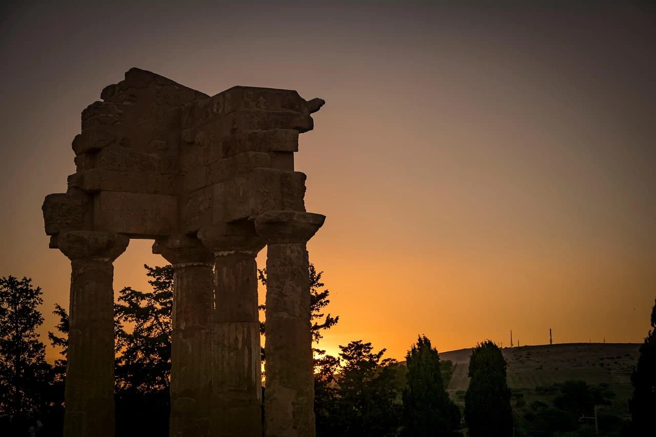 The silhouetted ruins of a Greek temple stand on a hill at sunset, with a dramatic, colorful sky.