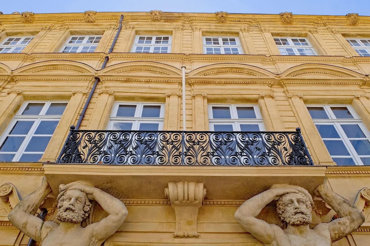 The intricate facade of a historic building is supported by two statues of men, with a decorative iron balcony in the center.