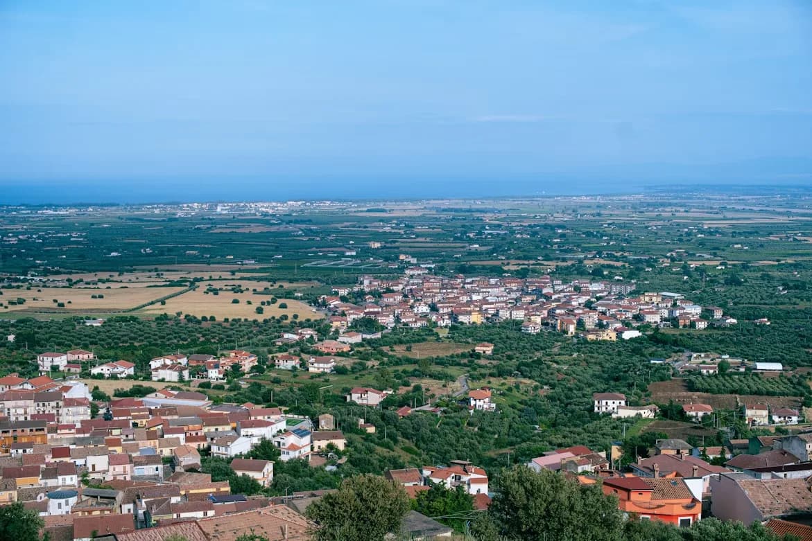 A panoramic view of a residential town shows a dense cluster of red rooftops and buildings surrounded by green fields and the sea in the distance.