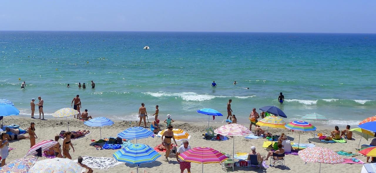 A bustling beach is filled with people and colorful umbrellas, with the blue sea and a clear horizon in the background.