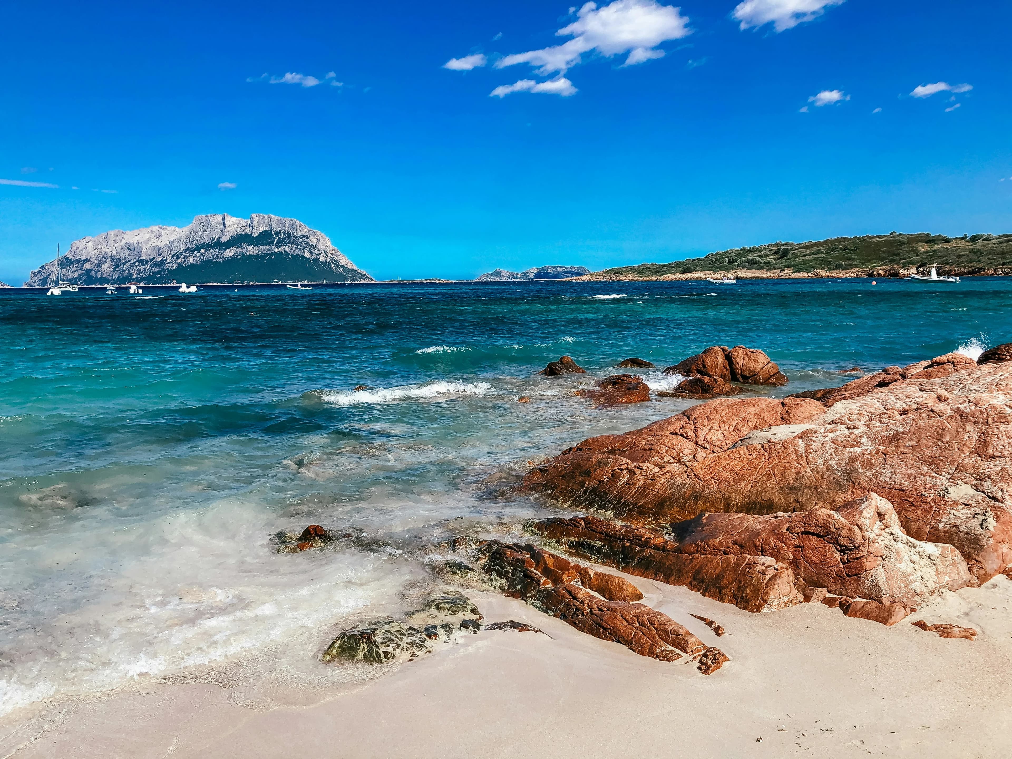 A picturesque beach with turquoise water, a white sandy shore, and large red rocks leading into the water, with a small mountain in the distance.