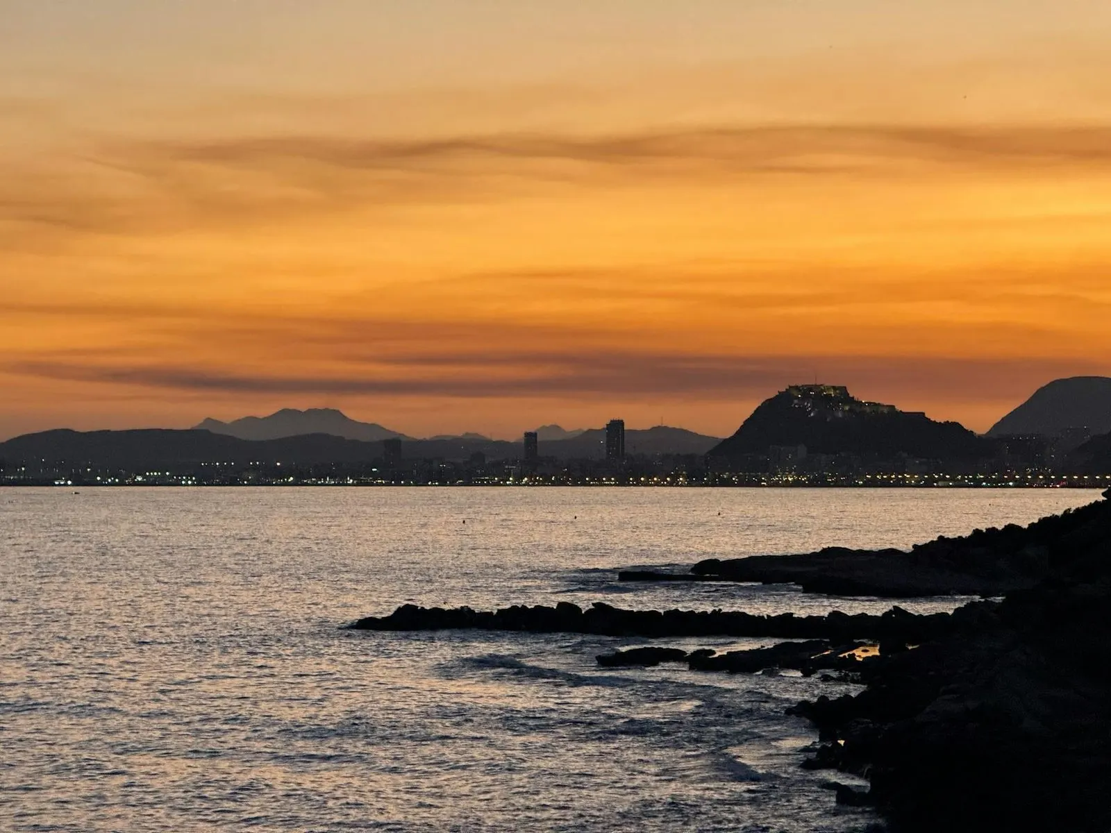 A beautiful golden sunset casts a warm glow over the sea, with a mountain and the lights of a city visible in the distance.