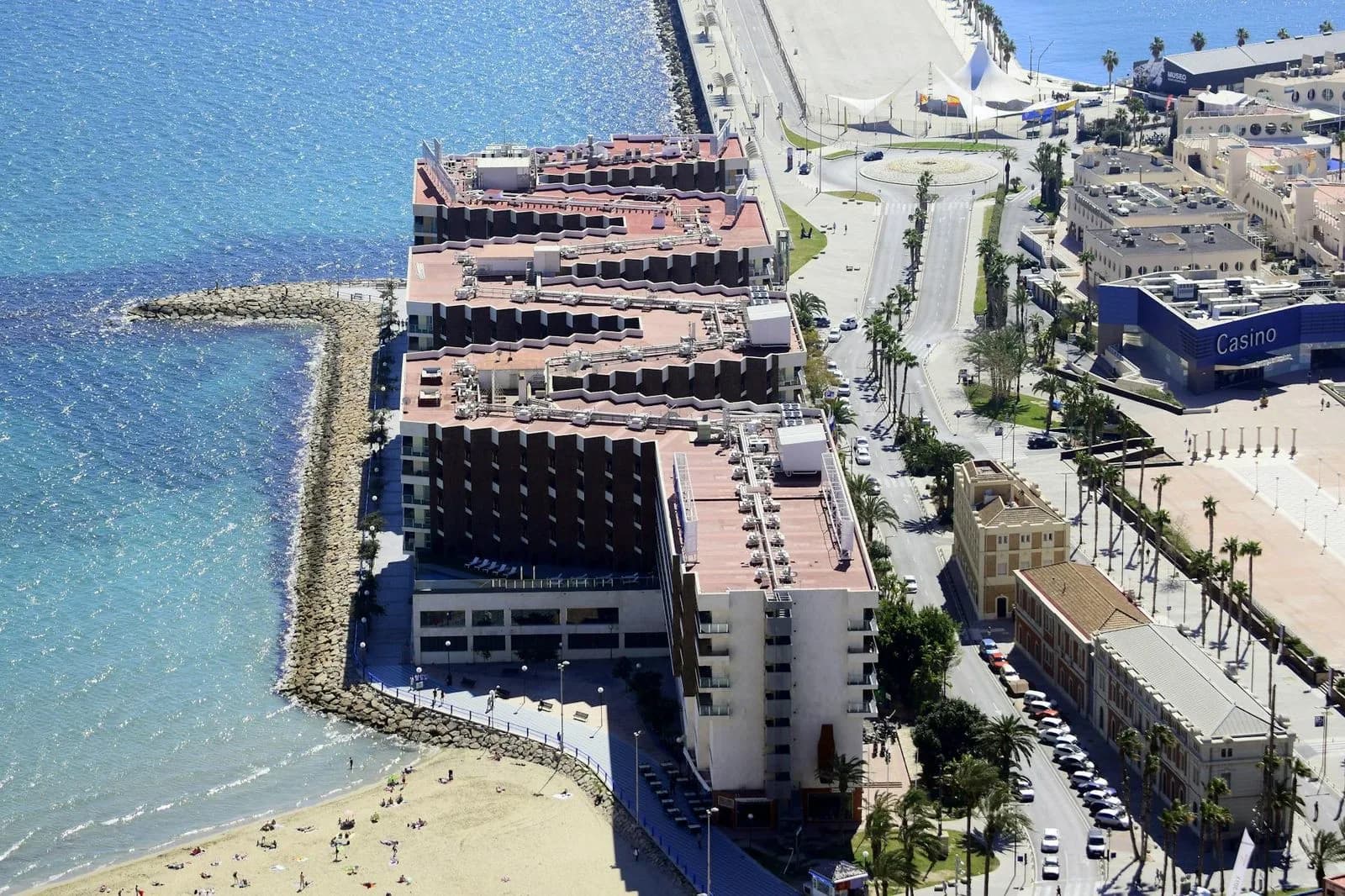 A high-angle view captures a modern hotel building on the waterfront, with a breakwater and the sea in the background.
