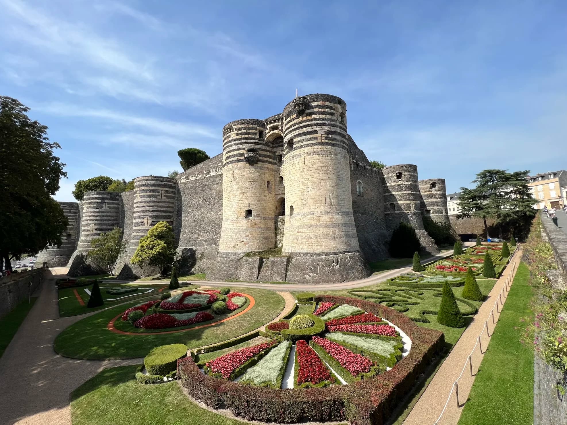 A panoramic view of the majestic Château d'Angers shows the fortress surrounded by beautiful, patterned gardens under a blue sky.
