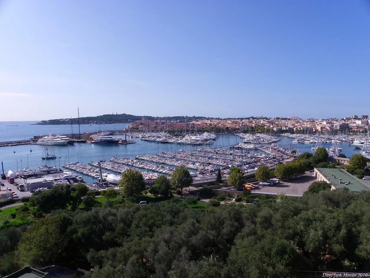 An aerial view captures the dense concentration of sailboats and yachts in the harbor, with a historic fortress on a hill in the background.