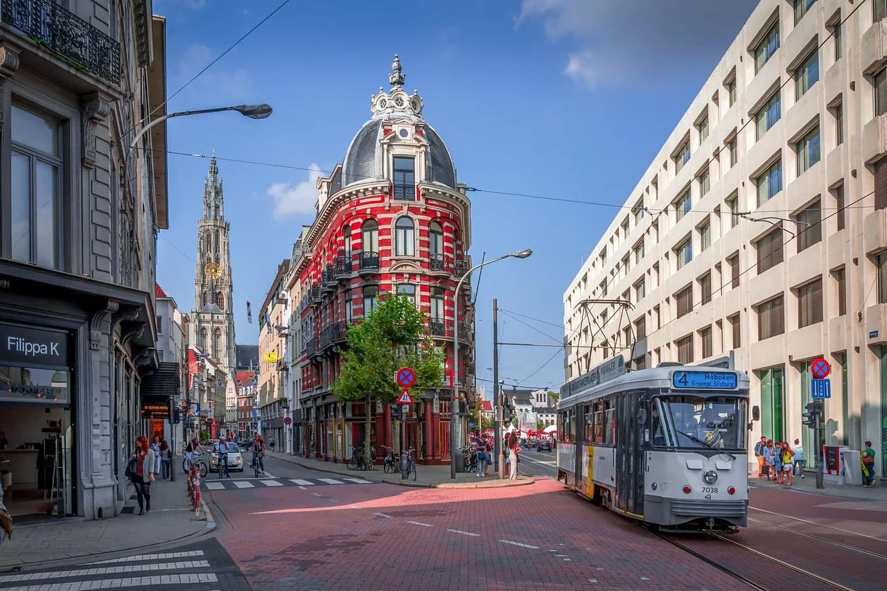 A tram travels along a city street lined with traditional and modern buildings, with the steeple of the Cathedral of Our Lady in the background.