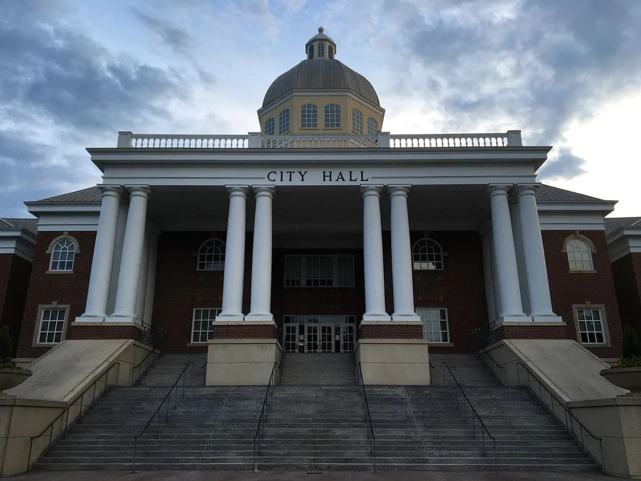 The grand facade of a city hall building, with its large dome and columns, stands against a cloudy sky.