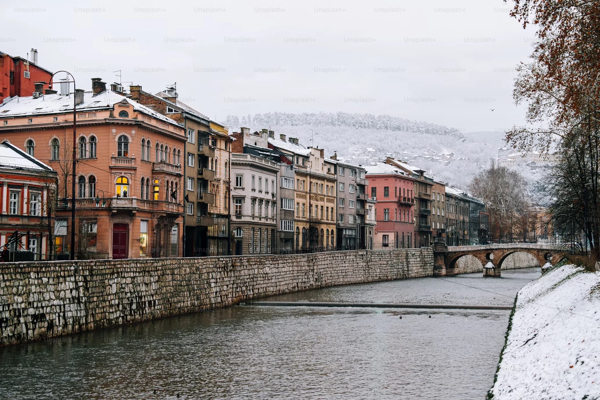 A quiet river flows through a city with a row of traditional houses and a bridge in the background, all covered in a light layer of snow.
