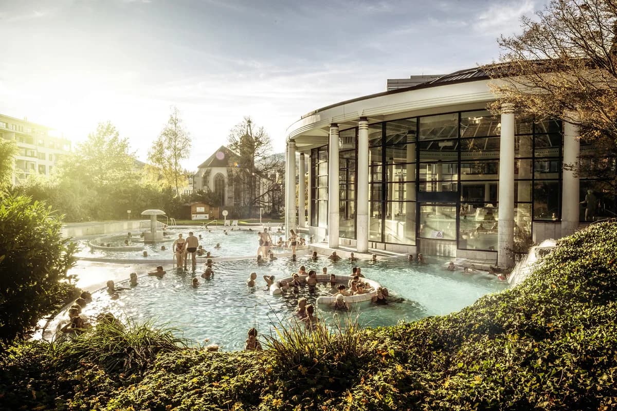 People relax and swim in a large, circular thermal pool in Baden-Baden, with a glass-enclosed building in the background.