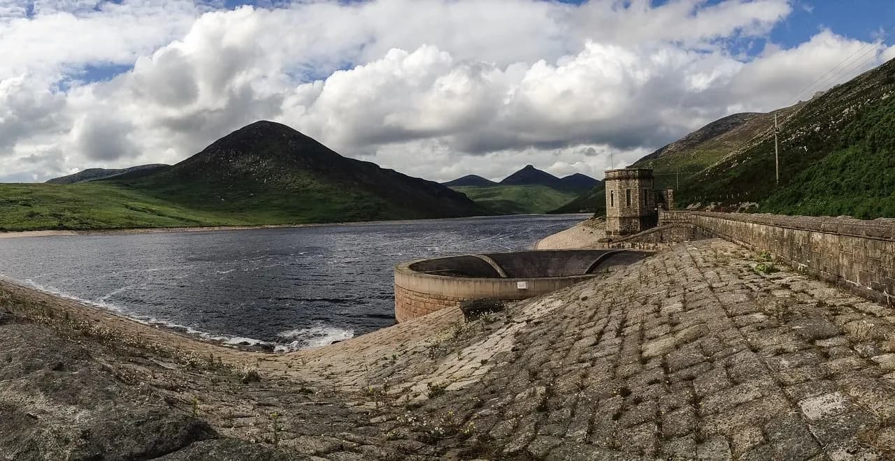 The majestic Mourne Mountains form the backdrop for a large reservoir, with a historic stone dam and a tranquil waterway in the foreground.