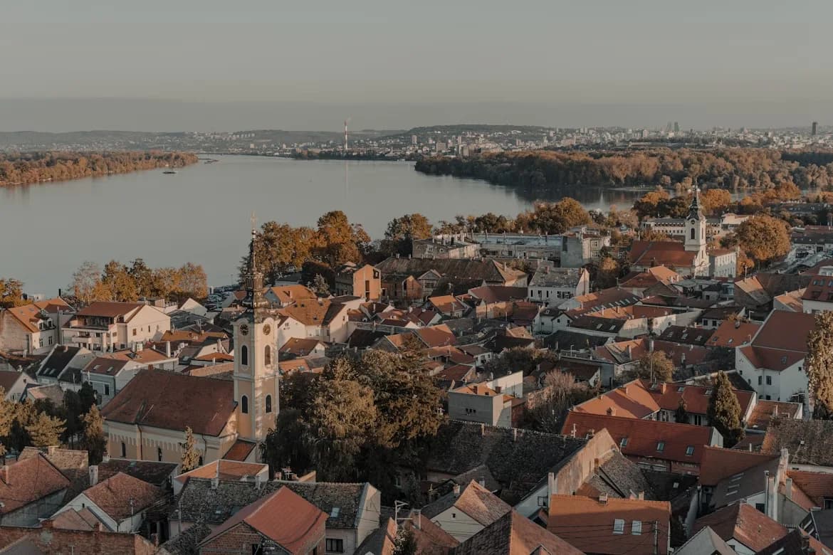 A high-angle view captures the red rooftops and church spires of the historic Zemun district, with the Danube River and the city of Belgrade in the distance.
