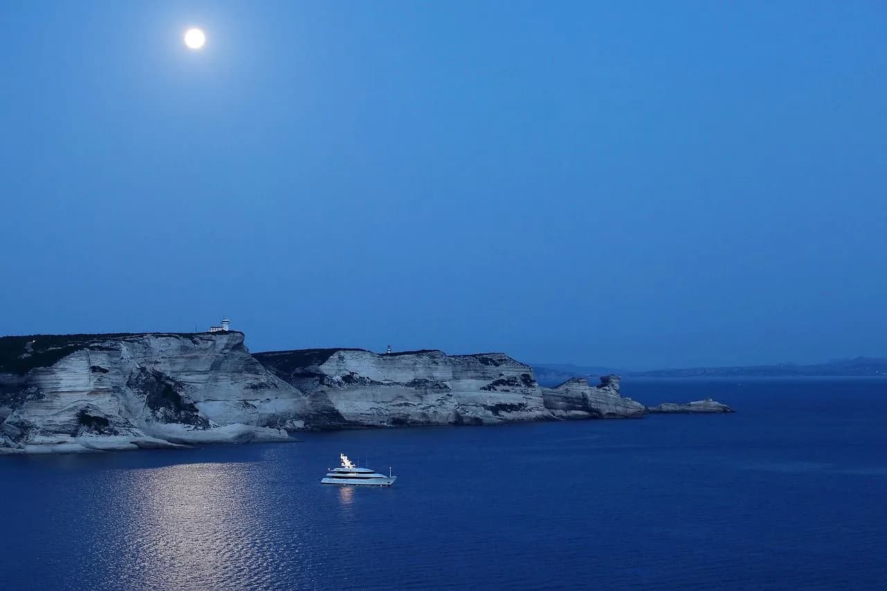 A luxury yacht is anchored in the calm blue sea under a full moon, with the dramatic white cliffs of Bonifacio illuminated in the distance.