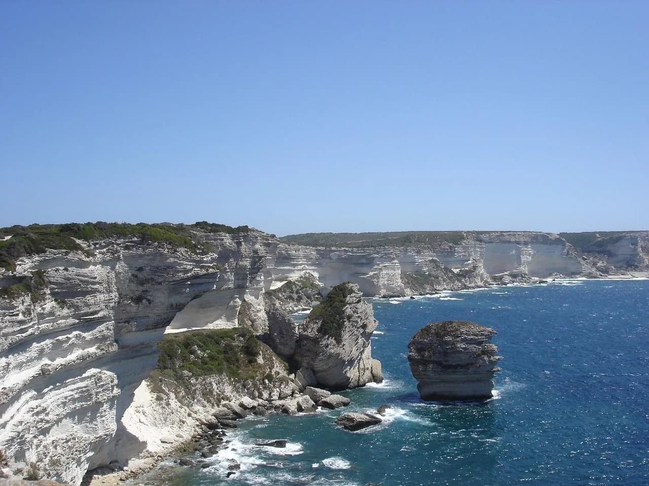 The dramatic white cliffs of Bonifacio plunge into the sea, with a few large rock formations rising from the water.
