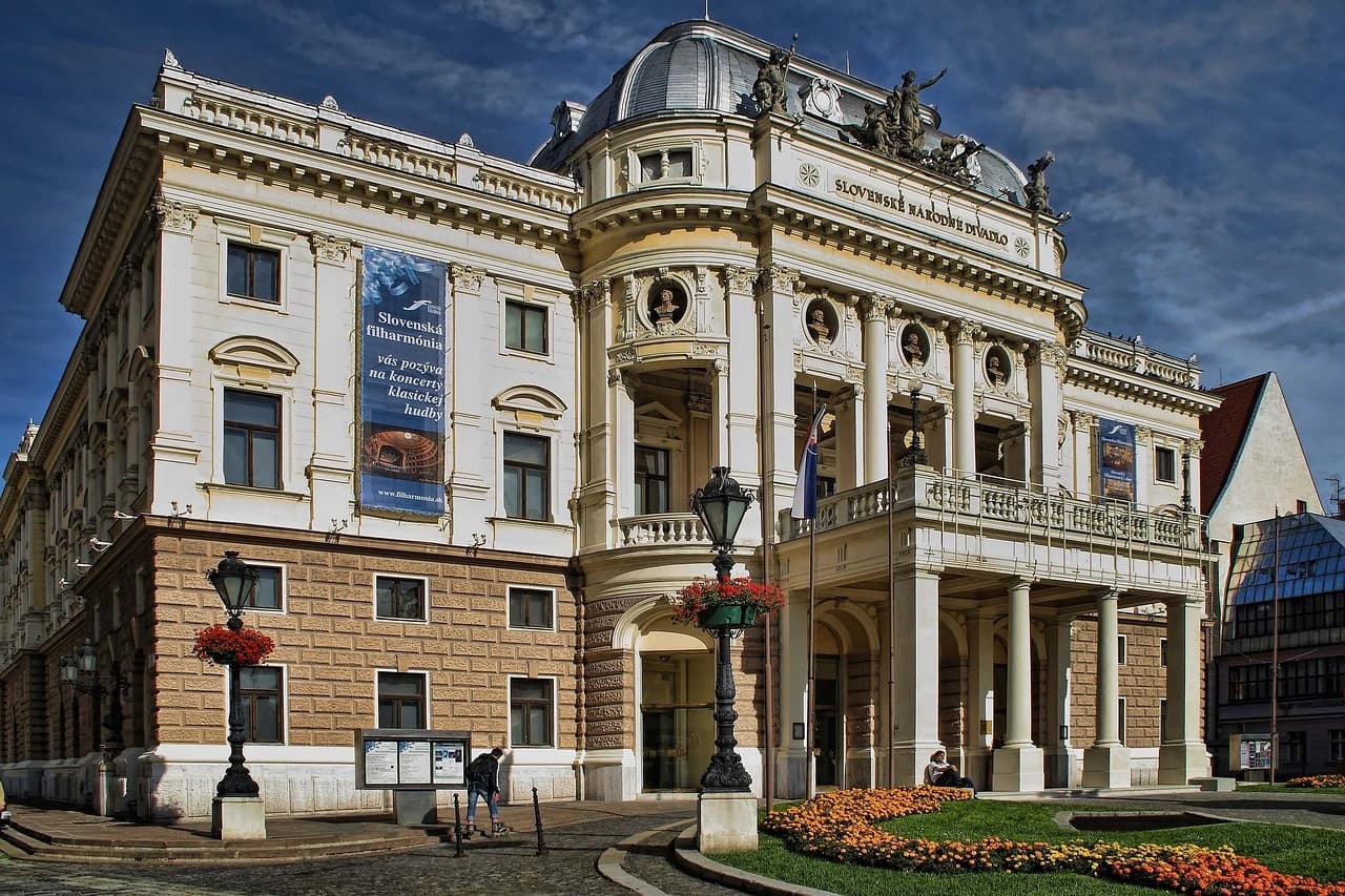 The elegant facade of the Slovak National Theatre building, with its grand columns and ornate roof, stands against a cloudy blue sky.