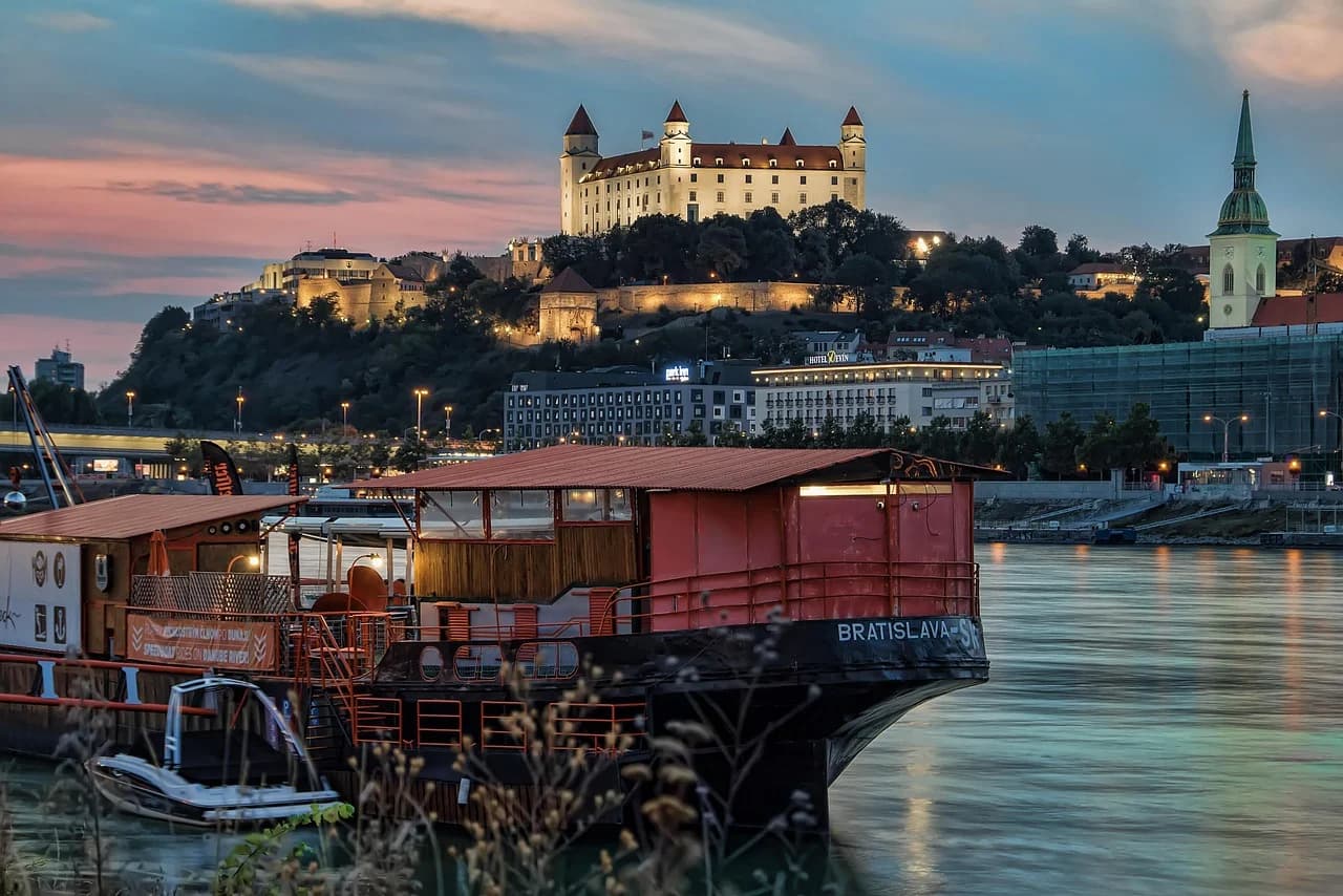 A historic castle is illuminated on a hill at dusk, with the lights of the city and a riverboat in the foreground.
