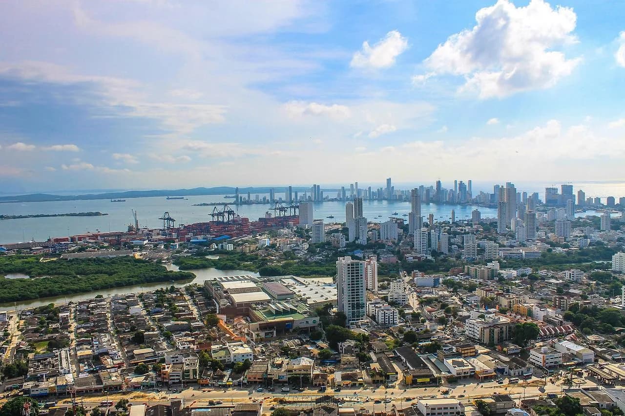 An aerial view captures the sprawling city of Cartagena, with its dense urban core, industrial port, and a modern high-rise skyline in the distance.