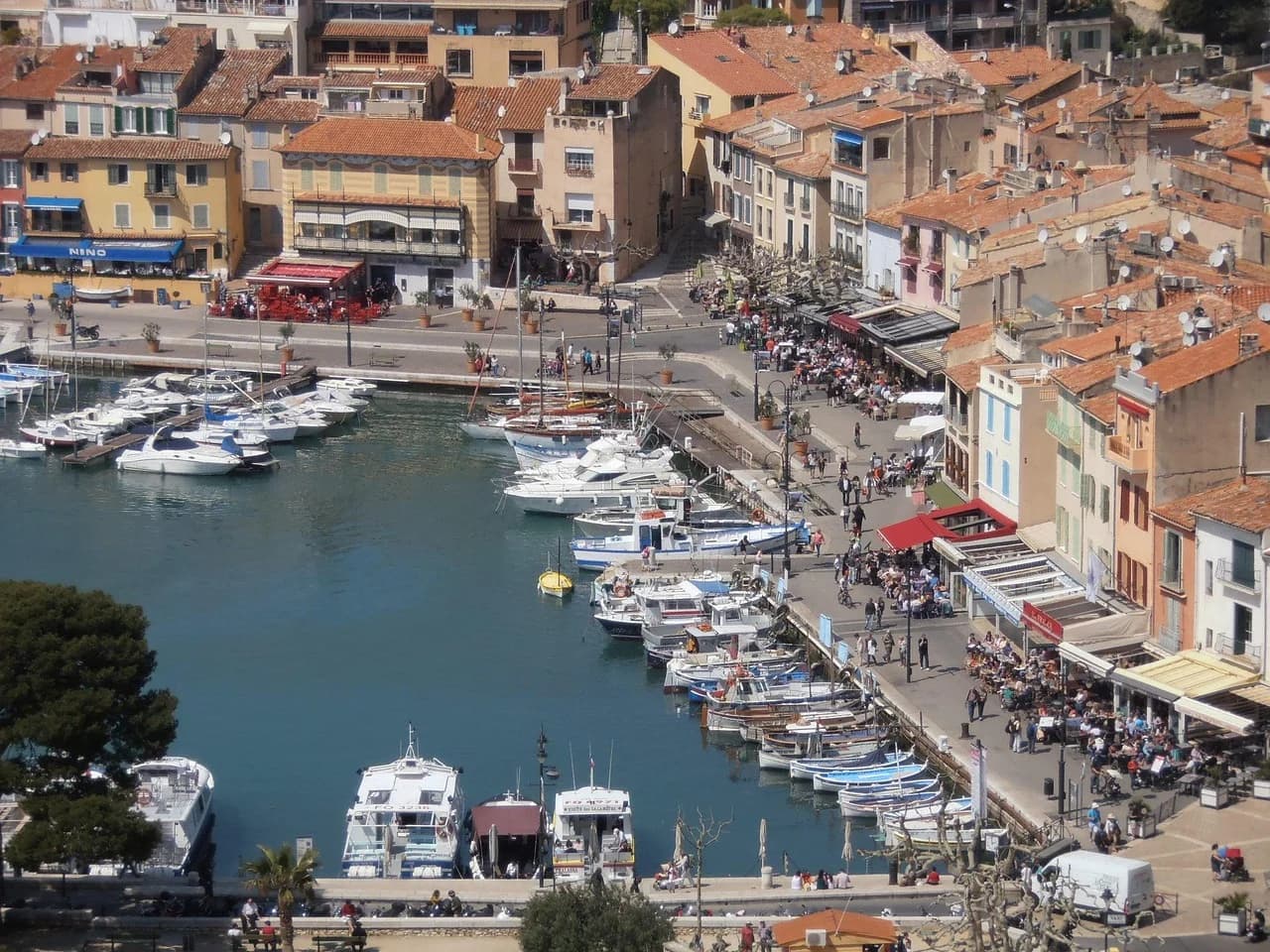 A high-angle view captures the lively port of Cassis, with numerous boats moored and people dining at the waterfront restaurants.