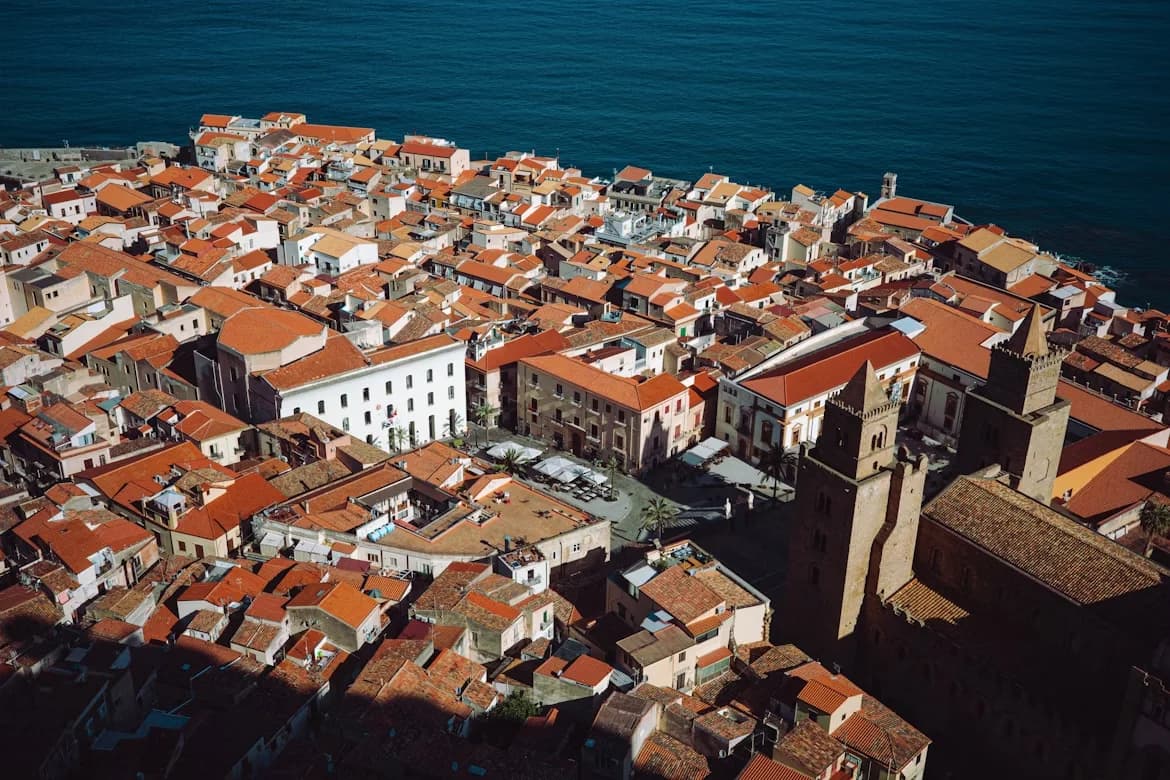 An aerial view shows the historic Cefalù Cathedral and the town's dense red-tiled rooftops, with the sea in the background.