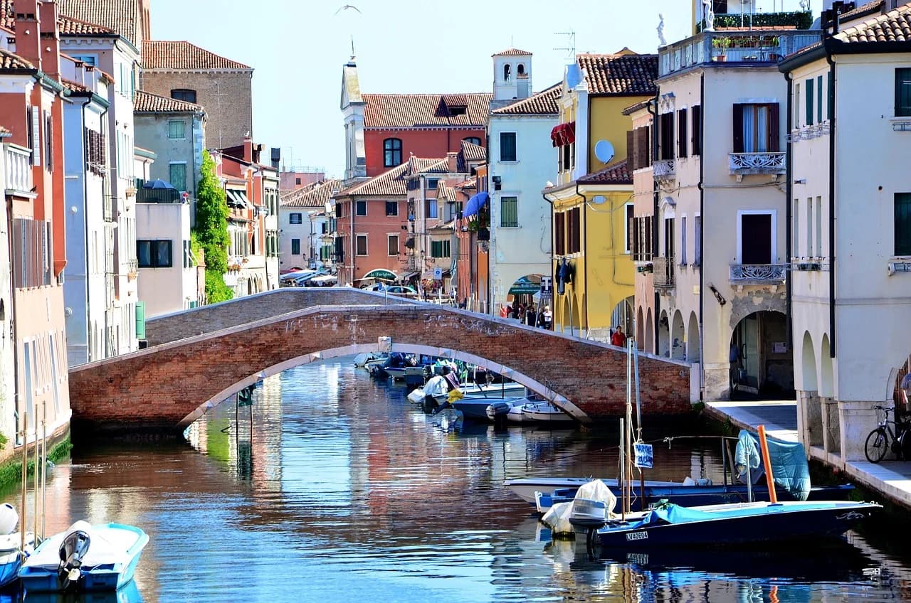 A quaint brick bridge spans a canal in Chioggia, with colorful, traditional buildings lining the waterway.