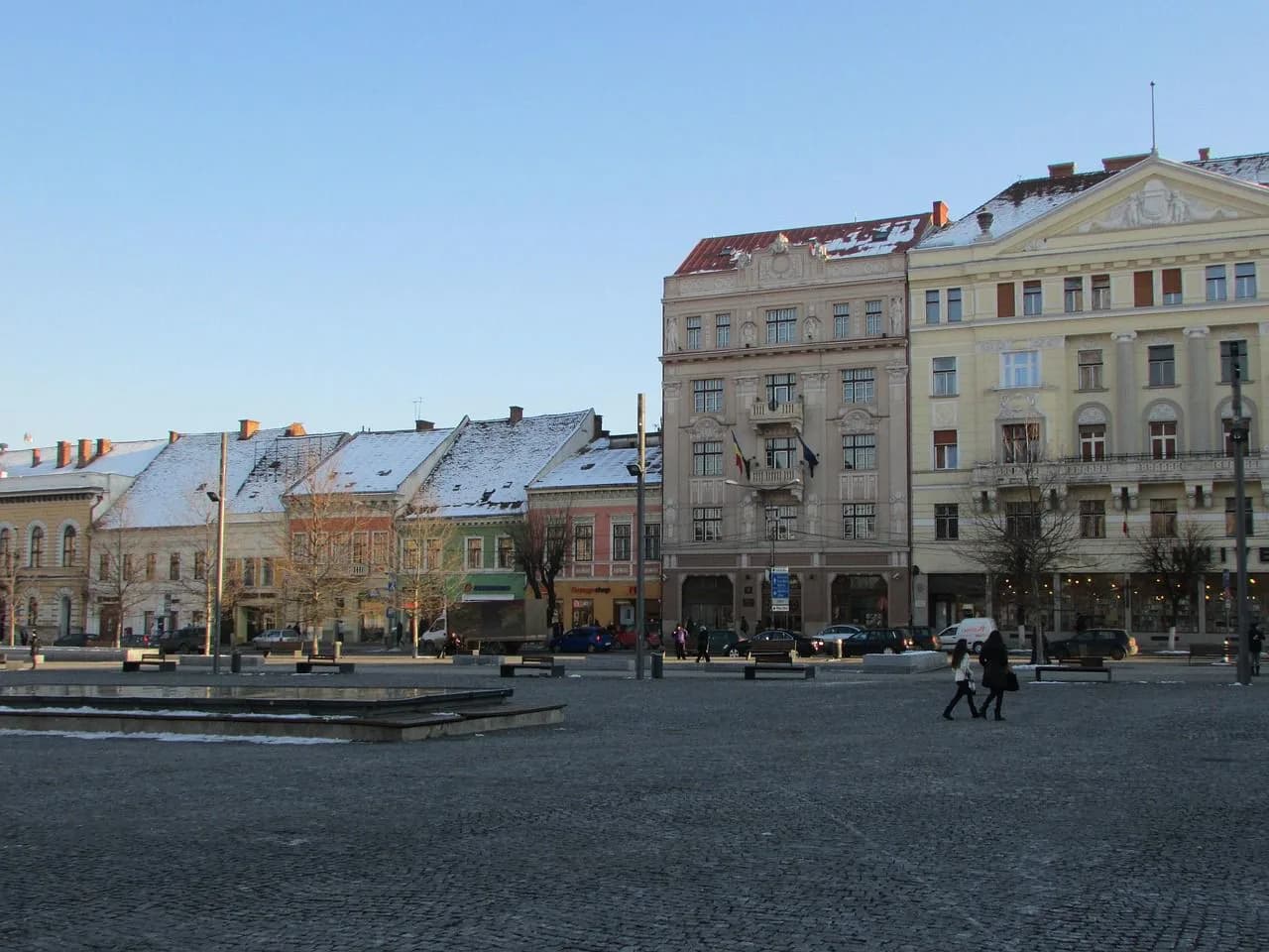 The city square of Cluj-Napoca is lightly covered in snow, with historic buildings and leafless trees under a clear winter sky.