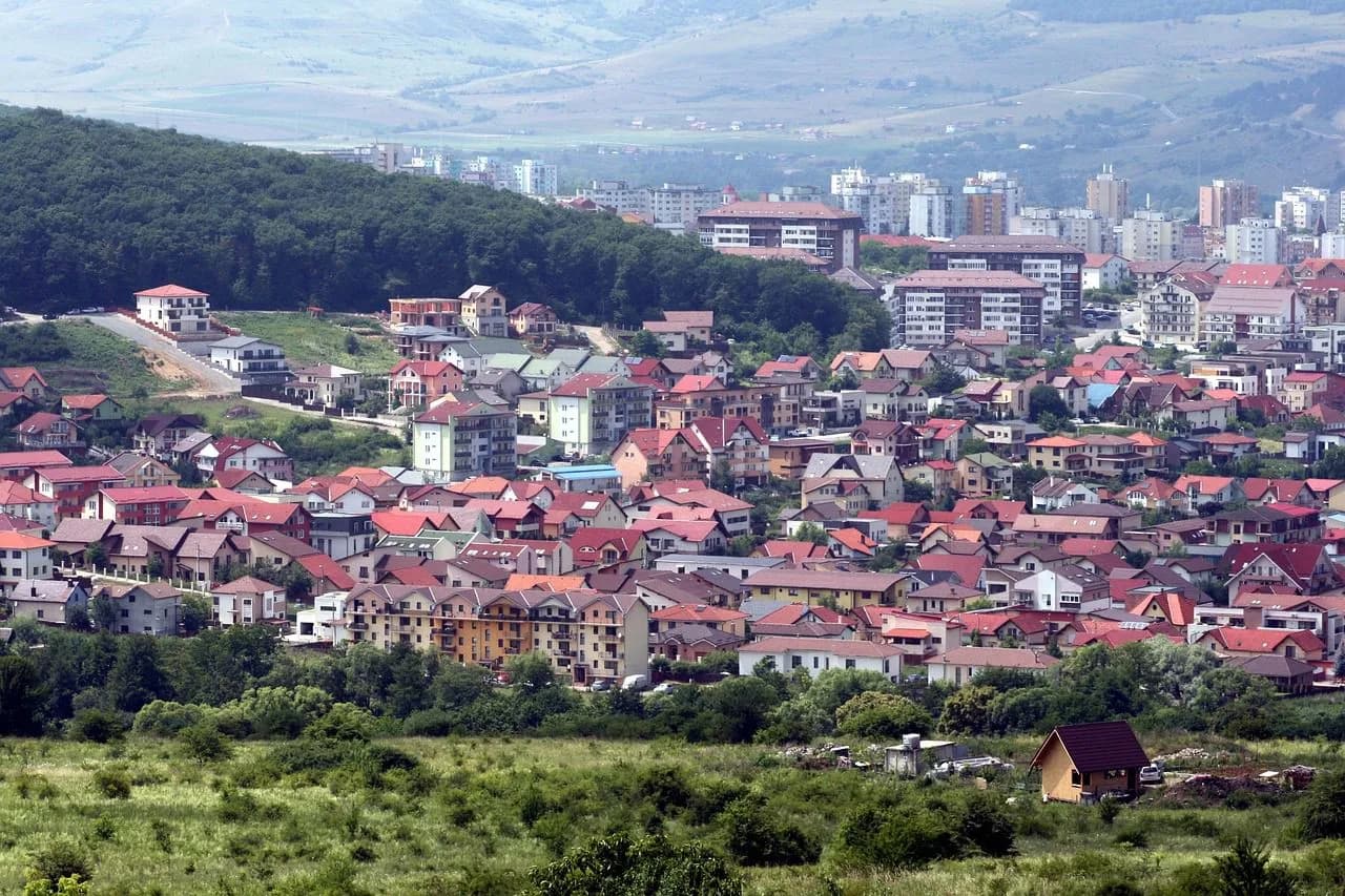 A panoramic view of Cluj-Napoca shows a mix of residential and modern buildings nestled in a valley with rolling green hills in the distance.