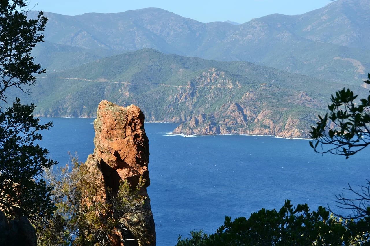 A rugged, red-rock formation stands in the foreground, providing a frame for the deep blue sea and the mountainous coastline in the distance.