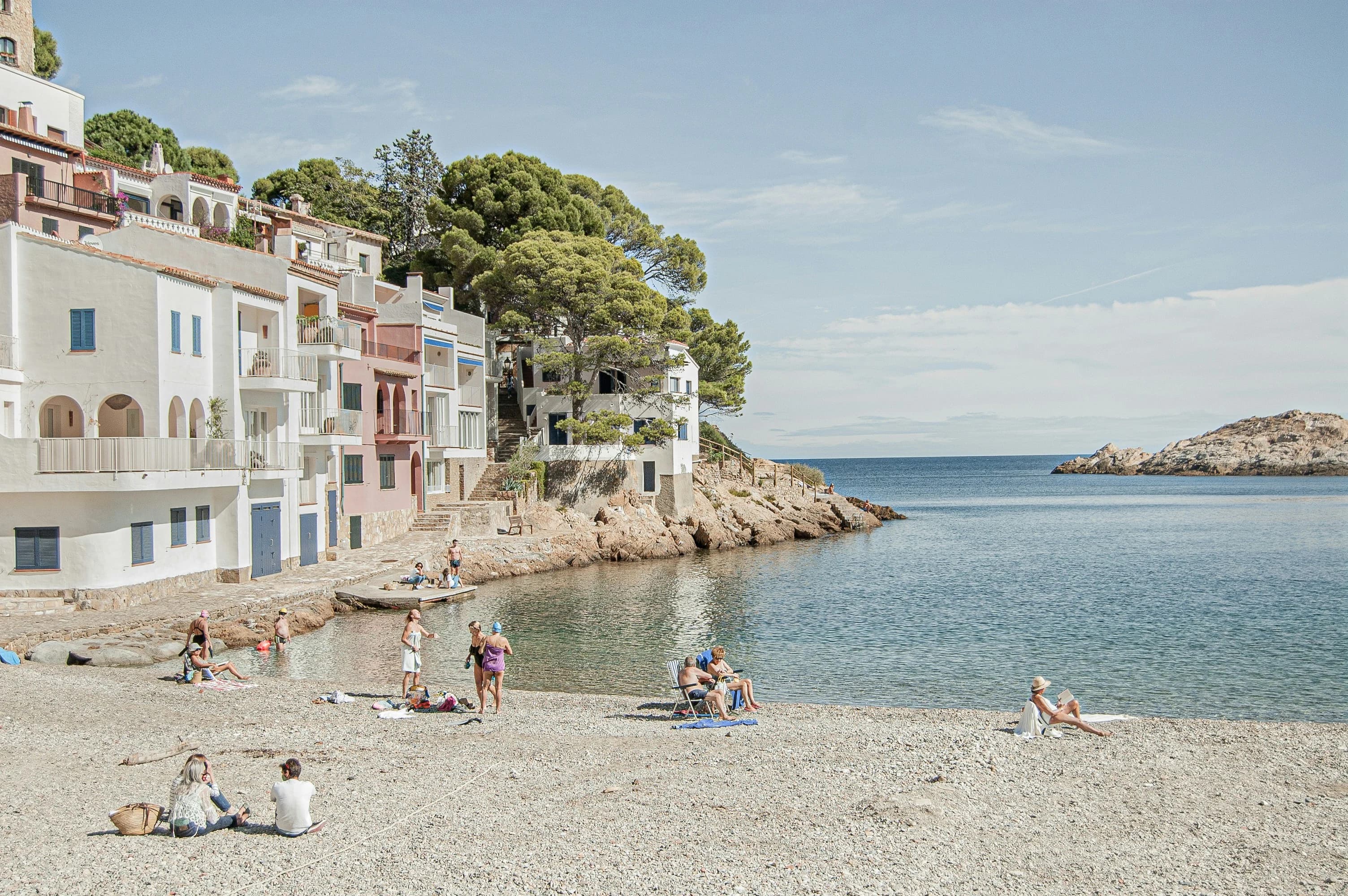 A group of people relax on a pebble beach, with white buildings and green trees built into the rocky coastline.