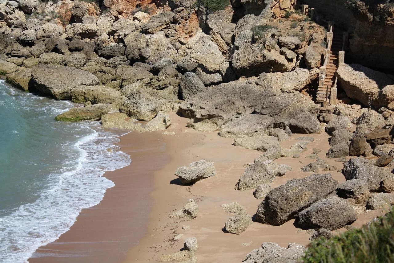 A secluded sandy beach is framed by large rocks and small waves, with a set of wooden stairs leading down to the shore.