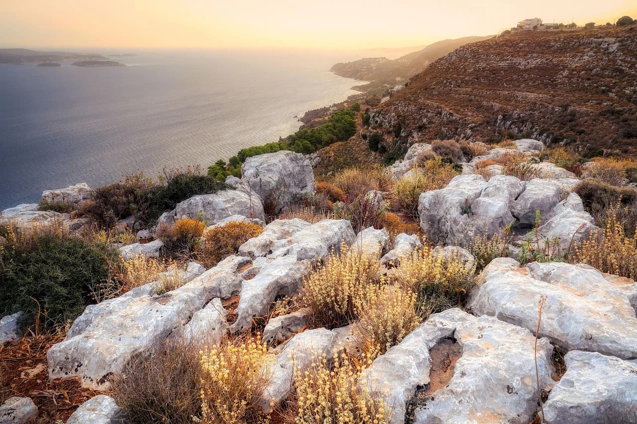 A scenic road winds along the rugged, rocky coastline of Crete at sunset, with the calm sea below and distant hills.