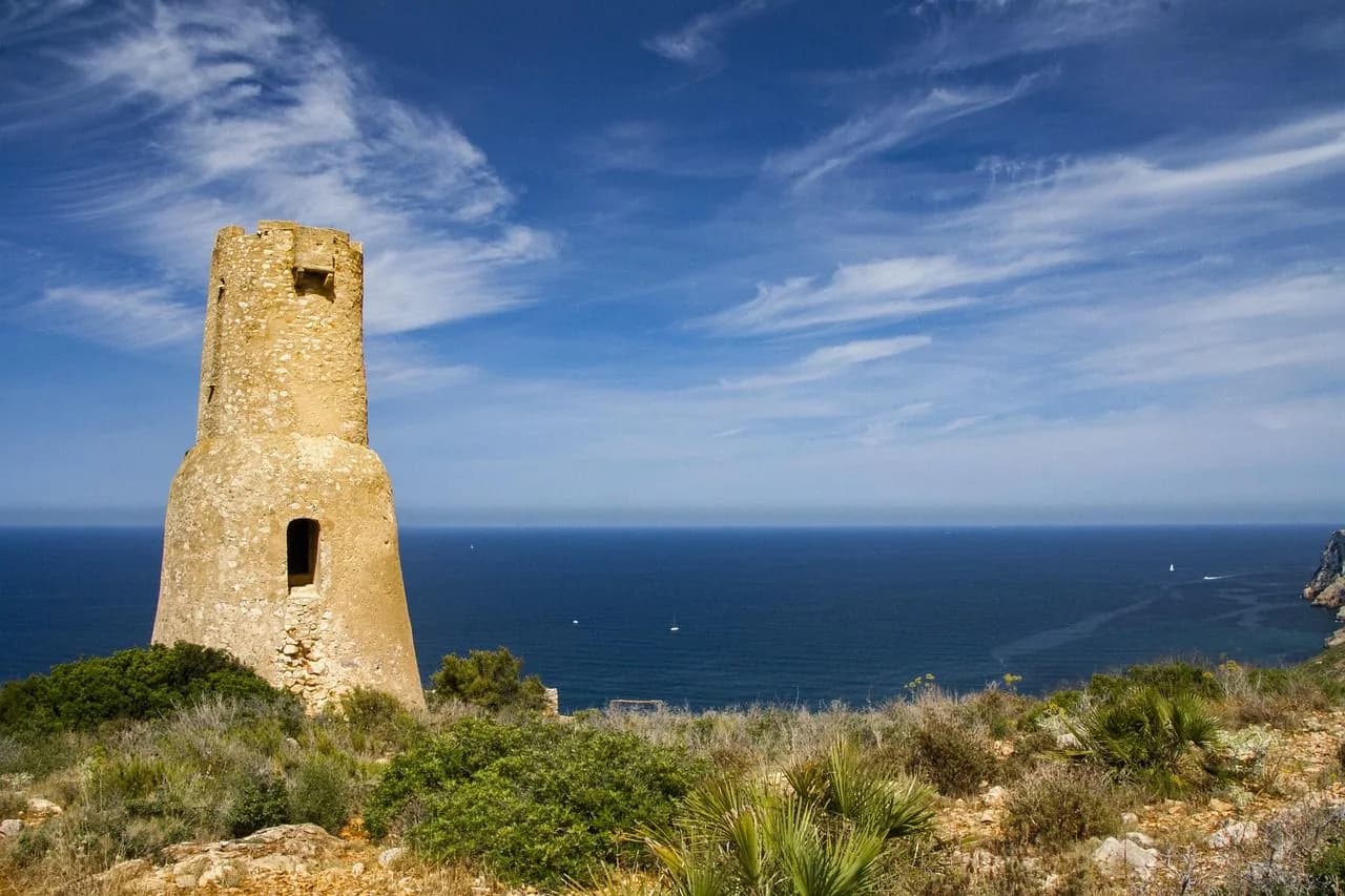 An ancient stone watchtower stands on a cliffside, overlooking the vast, open sea and a few sailboats in the distance.
