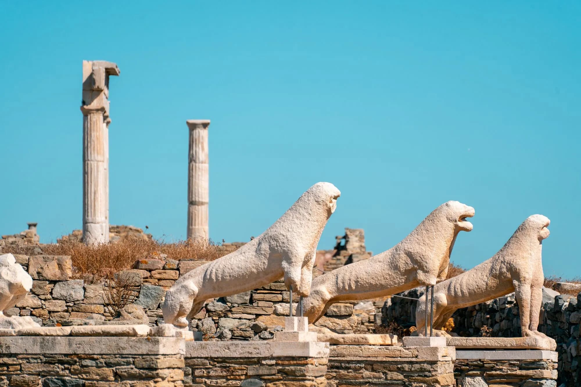 The famous marble lion statues of Delos are viewed from the side, their smooth forms contrasting with the rugged stone walls.