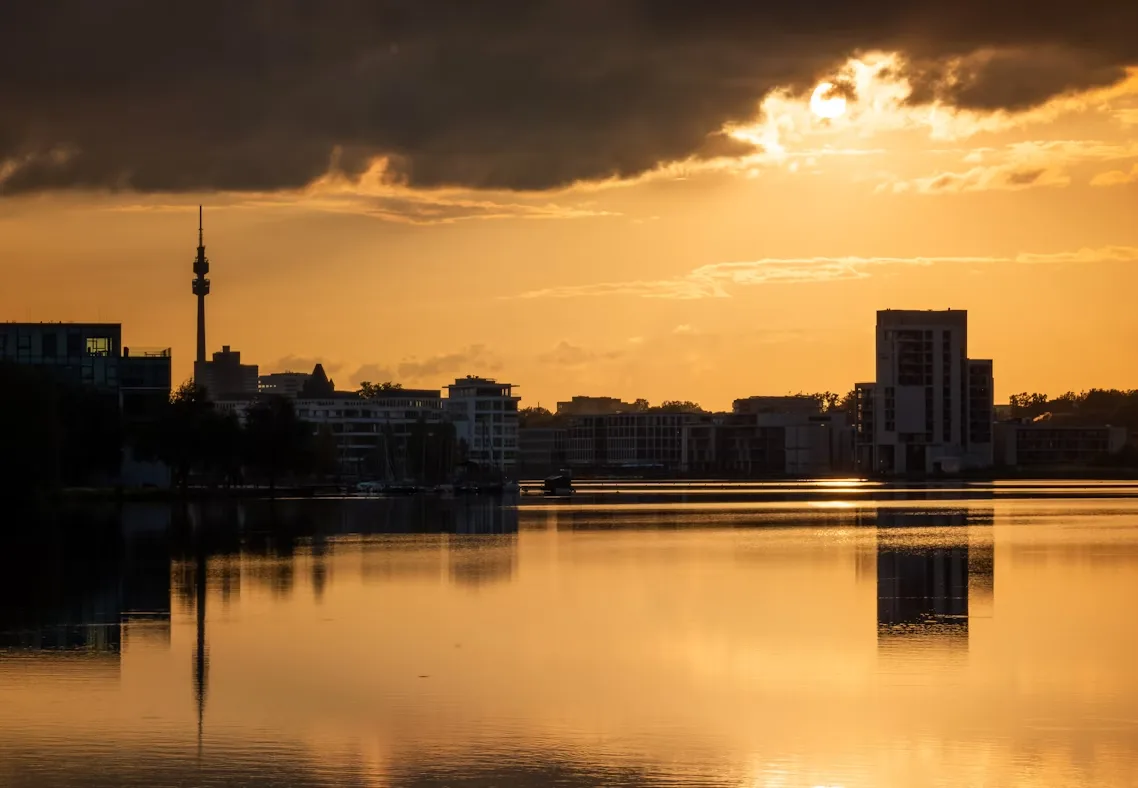 The Dortmund skyline, with the television tower and modern buildings, is silhouetted against a vibrant golden sunset, reflected in a tranquil lake.