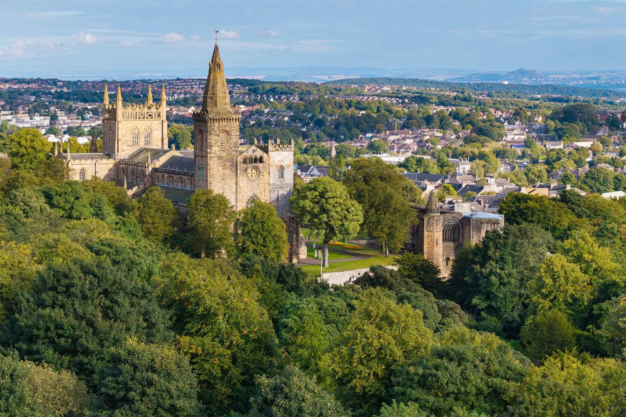 An aerial view captures the historic Dunfermline Abbey surrounded by a lush green landscape, with the city in the distance.
