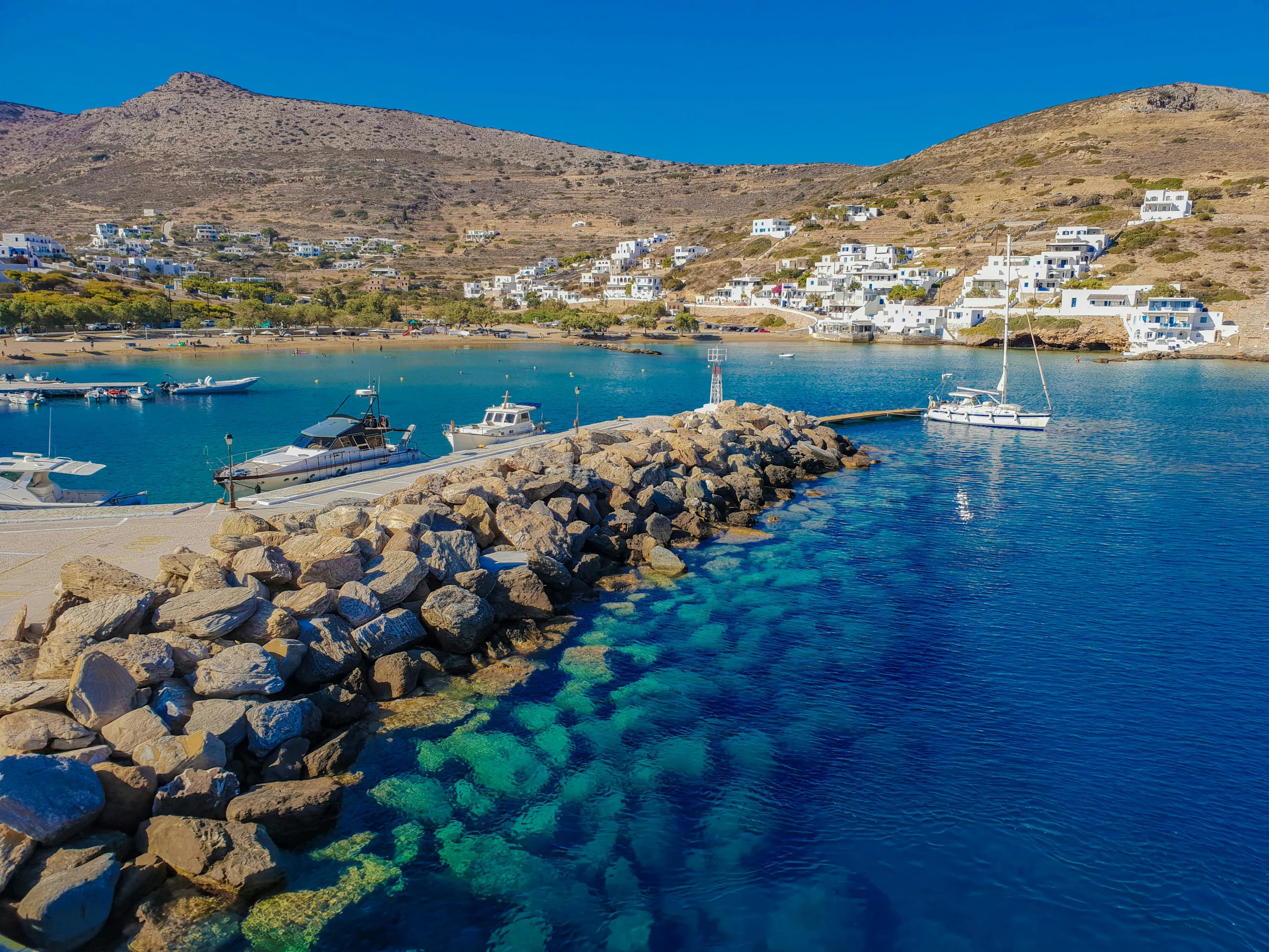 The serene harbor of Karavostasis is filled with boats, with the traditional white Cycladic buildings of the town built into the hillside.