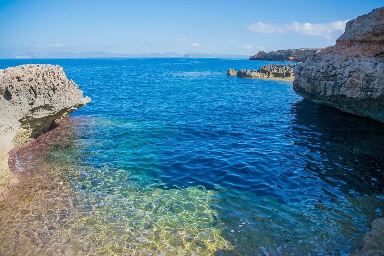 The shallow, crystal-clear turquoise waters of Formentera's coastline are framed by a rugged, rocky landscape.