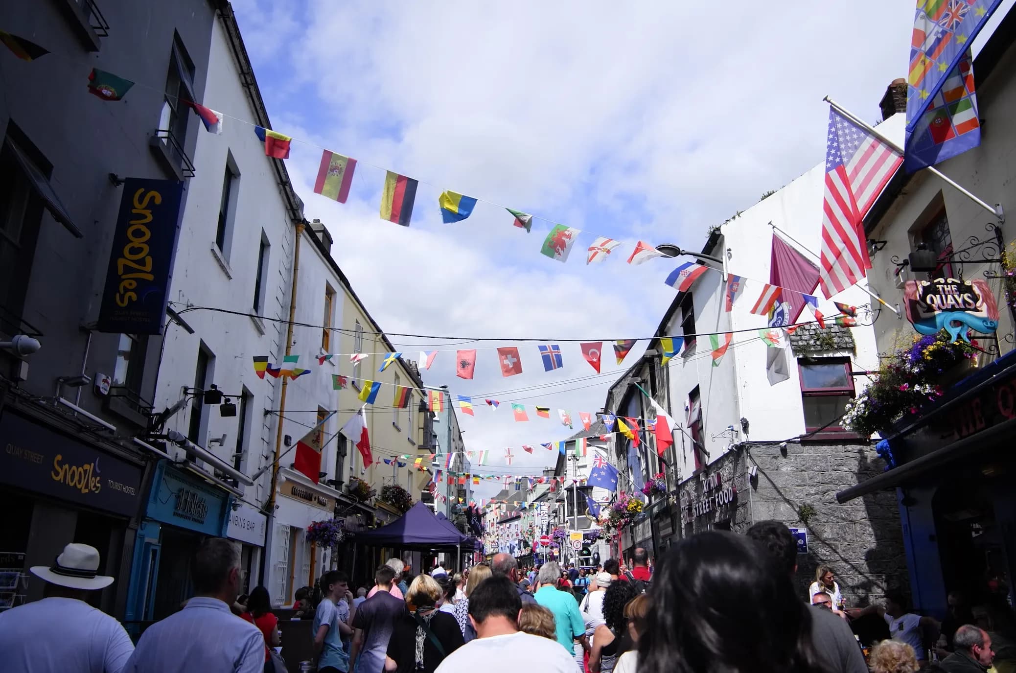 A street in Galway is decorated with flags from various nations, creating a festive atmosphere as people walk below.
