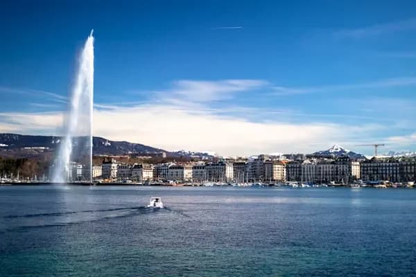 The Jet d'Eau, Geneva's iconic fountain, shoots water high into the air on a sunny day with the city and mountains in the background.