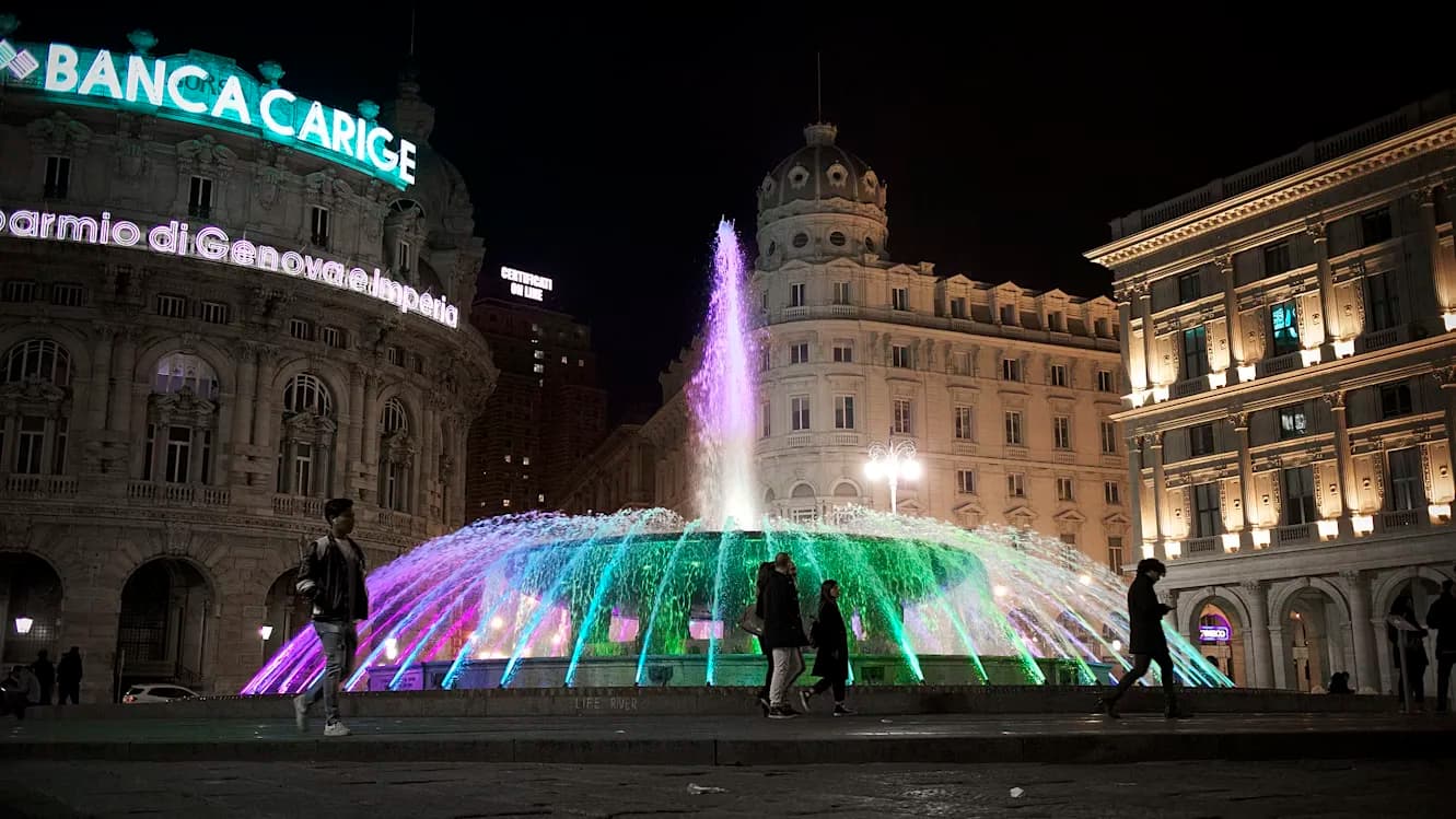 A brightly lit, multicolored fountain is the centerpiece of the Piazza de Ferrari, surrounded by historic buildings and people walking by at night.
