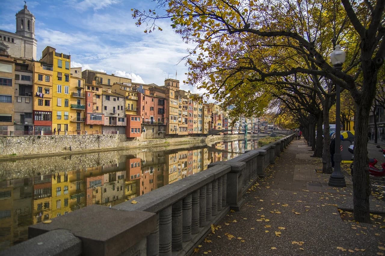The colorful, medieval buildings of Girona line the Onyar River, with a tree-lined promenade creating a beautiful autumn scene.